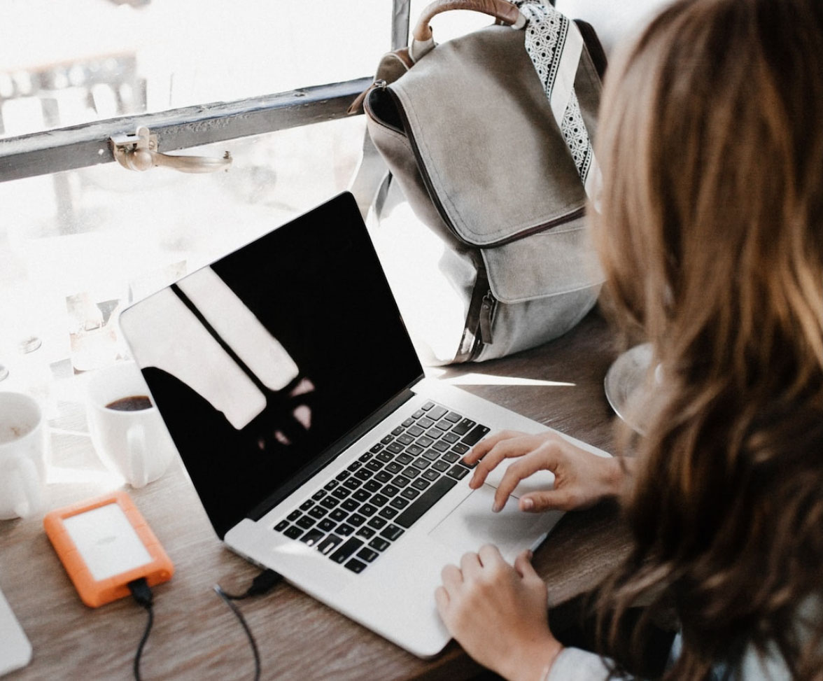 Person working on a laptop at a wooden counter, with an external hard drive, coffee and backpack