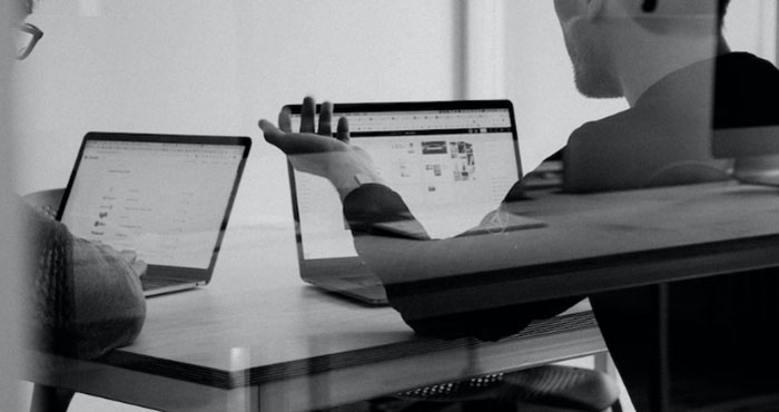 Two people working at laptops across a shared desk, seen through glass.