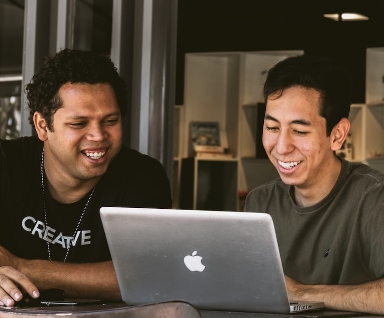 Two smiling colleagues looking at a laptop screen together in an open office.